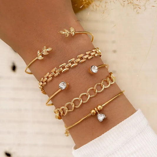 Poignet avec cinq bracelets dorés superposés, comprenant des motifs de feuilles, des maillons et des perles, ornés de cristaux.